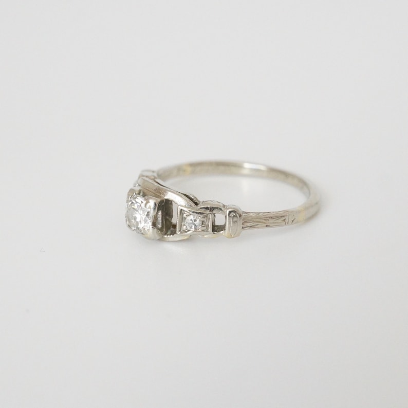 May include: A silver engagement ring featuring a single round diamond and two smaller diamonds set on the band. The band is engraved with a delicate pattern.