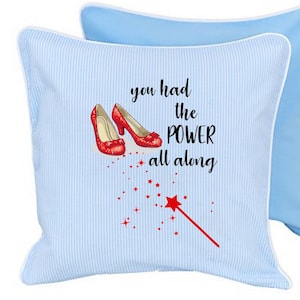 Peut inclure: Un coussin décoratif bleu clair à rayures et bordure blanche. Le coussin présente une illustration de chaussures rouges scintillantes, une baguette étoile rouge et le texte "you had the POWER all along."