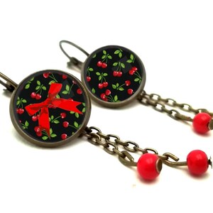 May include: A pair of dangle earrings with a black background and red cherries and a red bow. The earrings have a bronze chain with red beads.