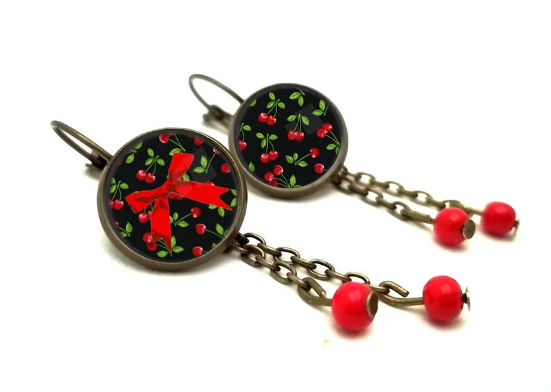 May include: A pair of dangle earrings with a black background and red cherries and a red bow. The earrings have a bronze chain with red beads.
