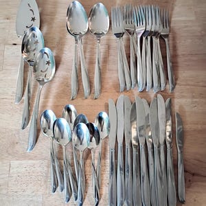 May include: A collection of stainless steel silverware, including spoons, forks, knives, and serving utensils. The silverware has a shiny finish and a decorative starburst design on the handles. The set is arranged on a wooden surface.