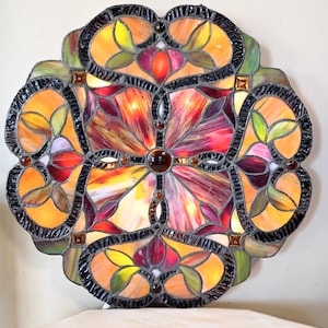 May include: A colorful, round stained glass lamp with a floral design. The lamp features a mix of orange, red, yellow, and green glass pieces, outlined with black. The center has a large, dark brown glass piece. The lamp is designed to be mounted on a wall or ceiling.