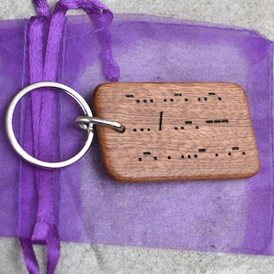 May include: A wooden keychain with a silver keyring, engraved with Morse code. The keychain is rectangular with rounded edges and is resting on a purple mesh bag with a matching ribbon. The Morse code is visible on the wooden surface.