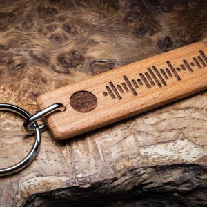 May include: A wooden keychain with a silver key ring. The keychain is rectangular and engraved with a black and white barcode-like pattern. The keychain is on a brown wooden surface.