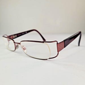 May include: Burgundy-colored eyeglasses with a rectangular frame and clear lenses. The temples are a darker shade of burgundy and feature a textured design. The glasses are set against a white background.