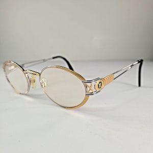 Puede incluir: Gafas de forma ovalada con montura dorada y plateada. Las monturas son doradas con un diseño decorativo y las patillas son plateadas con terminales negros. Las lentes son transparentes.