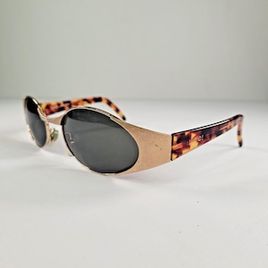 May include: Gold-coloured sunglasses with dark lenses and a tortoiseshell pattern on the arms. The sunglasses have a distinctive oval shape and a vintage style. The brand name is printed on the arm.