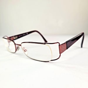 Puede incluir: Un par de gafas con montura de metal color granate oscuro y diseño semi al aire. Las varillas tienen un patrón texturizado y el puente conecta los cristales. Las plaquetas nasales son visibles y la estructura es elegante y funcional.
