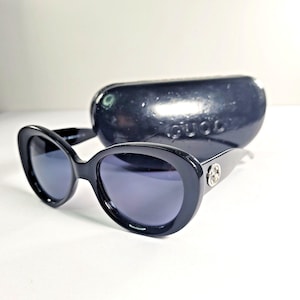 May include: Black oval-shaped sunglasses with dark tinted lenses and a silver-toned circular logo emblem on the temple. The sunglasses are accompanied by a matching black hard-shell protective case featuring the embossed Gucci brand name.