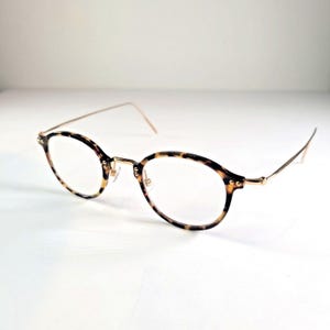 May include: Round eyeglasses featuring a tortoiseshell pattern frame with thin gold-tone metal temples and a matching bridge. The clear lenses are set within the circular rims, which are connected to the slender arms by small gold-tone hinges.