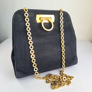 May include: A black suede handbag featuring a gold-tone metal Gancini closure on the front flap. The bag includes a long, decorative gold-tone chain strap composed of circular links that extends down from the top and pools at the base.