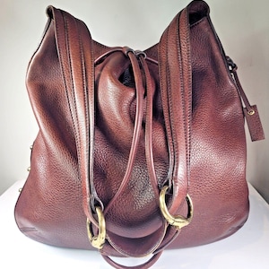 Puede incluir: Un bolso hobo de cuero granulado marrón oscuro con una silueta holgada, dos correas anchas para el hombro y anillos de metal dorado en la base de las correas. Una etiqueta de equipaje de cuero a juego cuelga del lado del bolso.