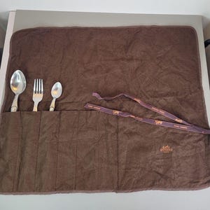 Może przedstawiać: Brązowy skórzany rolowany futerał na sztućce z sześcioma przegródkami. Futerał ma brązowy skórzany pasek z nadrukiem "HERMES PARIS".