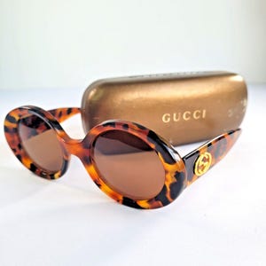 May include: Round sunglasses with a brown and black tortoiseshell pattern frame and dark brown lenses. The temples feature a gold-tone circular logo emblem. A matching metallic bronze-colored hard case with the word GUCCI printed in gold is included.