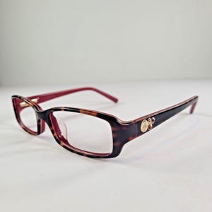 Könnte beinhalten: Rechteckige Brille mit Schildpattmuster auf den Fassungen und weinroten Bügeln. Die Brille hat ein goldfarbenes Logo und dekorative Akzente an den Bügeln. Die Gläser sind klar.