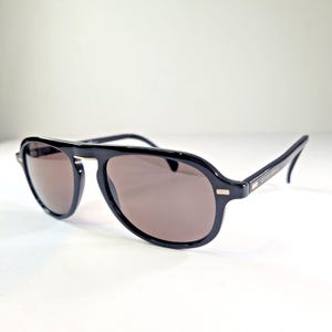 May include: Black aviator sunglasses with brown tinted lenses. The sunglasses have a thick, rounded frame and a double bridge. The arms are sleek and curve slightly at the ends. The lenses are a warm brown color.