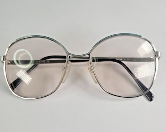 May include: Silver-framed eyeglasses with large, round lenses. The frames have a thin, metallic appearance with a subtle blue accent at the top. The lenses are a light pinkish hue. The glasses have a classic, vintage aesthetic.