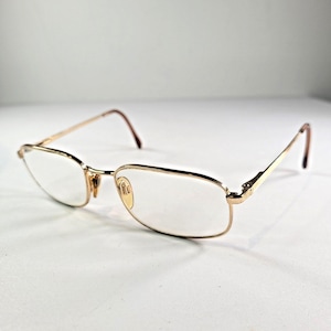 May include: Gold-rimmed spectacles with rectangular lenses. The glasses feature a slender frame and temples that curve at the ends. The lenses are clear, and the spectacles are set against a white background.