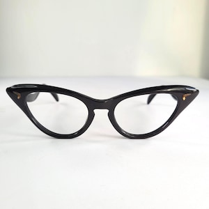 Puede incluir: Gafas negras de ojo de gato con un diseño retro. Las monturas son de color negro brillante con pequeños detalles dorados en las esquinas exteriores. Las lentes son transparentes, sobre un fondo blanco.