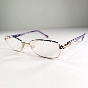 Może przedstawiać: Okulary z oprawkami w kolorze złotym i fioletowymi zausznikami. Soczewki są przezroczyste, a oprawki mają dekoracyjny wzór w pobliżu zawiasów. Zauszniki są w jednolitym fioletowym kolorze.