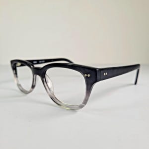 May include: Black cat-eye style eyeglasses with a gradient lens. The frames are black with two small silver rivets on the front and side arms. The lenses transition from clear to a smoky gray color. The glasses are set against a white background.