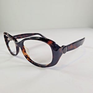 May include: Tortoiseshell eyeglasses with a cat-eye frame. The glasses feature a dark brown and amber pattern, with a small emblem on the temples. The lenses are clear.