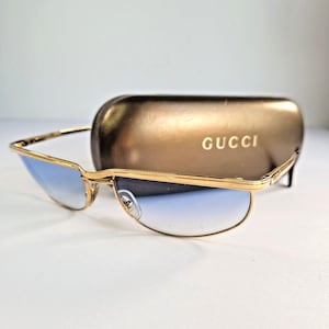 May include: Gold-tone metal sunglasses with blue gradient lenses and a thin frame. Behind the sunglasses sits a metallic bronze-colored hard case with the word GUCCI printed in gold lettering on the front surface of the protective eyewear case.