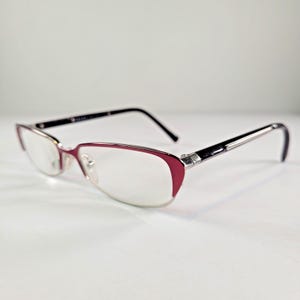 Op de afbeelding: Een bril met een bordeauxrode en zilveren montuur. De glazen zijn helder en de veren zijn zwart. De bril staat tegen een witte achtergrond.