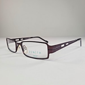May include: Rectangular eyeglasses with a dark brown frame. The lenses are clear, and the temples have a geometric design. The brand name "ZENITH" and the word "ZEST" are printed on the lens.