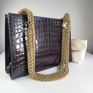 May include: A dark brown, rectangular handbag with a gold chain strap. The bag has a crocodile skin texture and a flap closure. A small, off-white pouch with the brand name is visible.