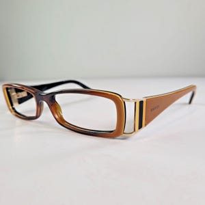 Op de afbeelding: Bruine en zwarte Gucci-bril met een rechthoekig montuur. De bril heeft een kleurenschema met een verloop, van donkerbruin naar lichtbruin. De veren zijn effen bruin met gouden accenten en het Gucci-logo.
