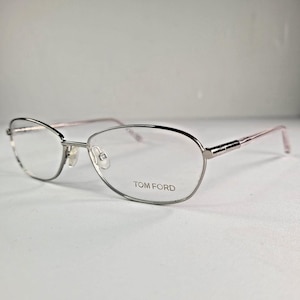 Może przedstawiać: Srebrne okulary Tom Ford z delikatną, owalną oprawką. Zausznik jest w kolorze jasnoróżowym, a logo Tom Ford jest widoczne na soczewce. Okulary mają nowoczesną, minimalistyczną estetykę.