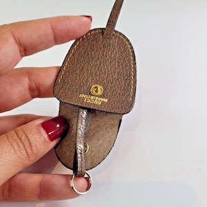 Puede incluir: Un llavero de cuero marrón con un llavero plateado. El llavero está grabado con un patrón texturizado y presenta un cierre de botón a presión dorado. El texto "GUCCI MADE IN ITALY" es visible.