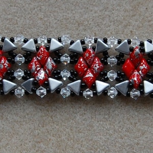 Puede incluir: Un brazalete de cuentas plateadas y negras con cuentas de forma de diamante rojas y blancas. El brazalete tiene un cierre de palanca plateado.