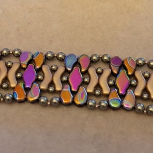 May include: Close-up of a bracelet with iridescent teardrop-shaped beads in shades of purple, orange, and blue. The design includes small, round gray beads and curved, light-colored beads.
