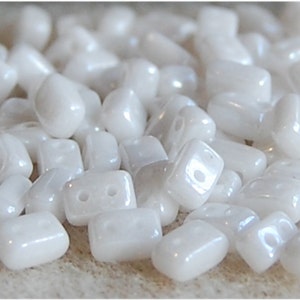 May include: A pile of white, opaque, rectangular glass beads with two holes in each bead. The beads have a slightly iridescent finish.