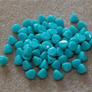 May include: A pile of turquoise blue glass beads in a teardrop shape with a pointed tip. The beads are arranged in a random pattern on a light brown surface.