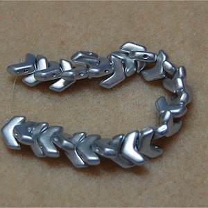 May include: A strand of silver-colored arrow-shaped beads. The beads are connected to form a continuous chain.