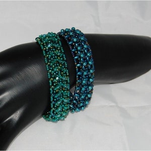 Pode incluir: Duas pulseiras de contas, uma verde e outra azul, em um bra&ccedil;o de manequim preto. As pulseiras s&atilde;o feitas de pequenas contas iridescentes.