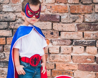May include: A young child dressed as a superhero wearing a blue cape, a red mask with a spider design, a red belt with a spider buckle, and blue jeans. The child is standing in front of a brick wall and holding a red shield with a black spider design.