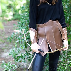 May include: A woman dressed in a black hooded tunic and brown leather skirt holds a wooden bow and arrow. She is wearing a brown leather headband with a white diamond shape on it. The background is a blurred green forest.