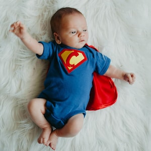 May include: A baby wearing a blue Superman costume with a red cape. The baby is lying on a white fluffy surface.