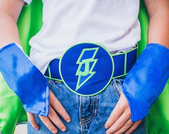 May include: A child wearing a green and blue superhero costume with a blue belt featuring a lightning bolt and the letter "J".
