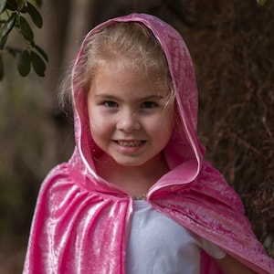 May include: A young girl wearing a pink velvet hooded cape. The cape has a satin trim and is gathered at the shoulders. The girl is smiling and looking at the camera.