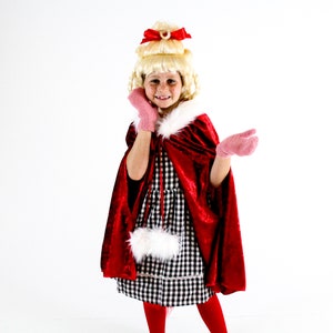 May include: A young girl wearing a red velvet cape with a white fur trim, a black and white gingham dress, and pink mittens. She has blonde hair styled in a bouffant with a red bow.