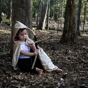 May include: A young person dressed in a hooded cloak holds a toy bow and arrow in a wooded area. The cloak is a light beige color with a brown hood. The bow and arrow are made of wood and have a black string. The person is sitting on the ground with their legs crossed. The background is a forest with trees and leaves.