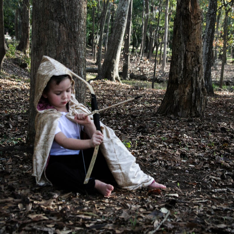 May include: A young person dressed in a hooded cloak holds a toy bow and arrow in a wooded area. The cloak is a light beige color with a brown hood. The bow and arrow are made of wood and have a black string. The person is sitting on the ground with their legs crossed. The background is a forest with trees and leaves.