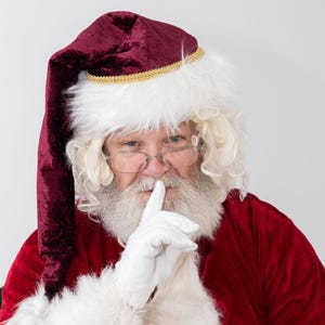 May include: A Santa Claus wearing a red velvet suit and a white fur-trimmed hat with a gold trim. He is holding his finger to his lips, as if to say 'shhh'.