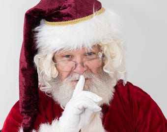 May include: A Santa Claus wearing a red velvet suit and a white fur-trimmed hat with a gold trim. He is holding his finger to his lips, as if to say 'shhh'.