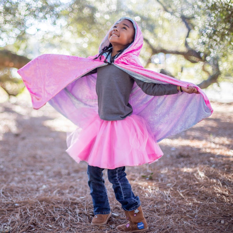 May include: A young person wearing a pink and blue hooded cape with a pink tulle skirt and brown boots. The cape has a sparkly iridescent finish.